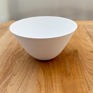 May include: A white ceramic bowl with a smooth, rounded interior and a slightly flared rim. The bowl is sitting on a wooden surface.
