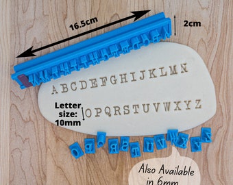 May include: A blue plastic alphabet stamp set with individual stamps for each letter of the alphabet. The stamps are 10mm in size and are arranged in a line on a white surface. The set also includes a ruler that measures 16.5 cm long. The text "Also Available in 6mm Size!" is written in a speech bubble.