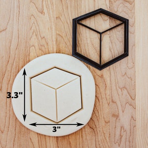 May include: A black plastic cookie cutter in the shape of a 3D cube. The cutter is 3 inches wide and 3.3 inches tall. The cutter is shown on a wooden surface with a sample cookie dough cutout.