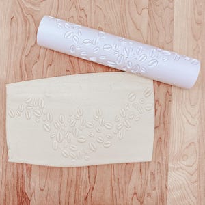 May include: A white rolling pin and a rectangular piece of dough, both embossed with a coffee bean pattern, rest on a wooden surface. The rolling pin is cylindrical, and the dough is light beige. The coffee bean design is repeated across both items.
