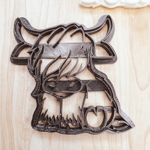 Puede incluir: Cortador de galletas de pl&aacute;stico negro con forma de vaca de las tierras altas con una melena esponjosa y una nariz en forma de coraz&oacute;n.