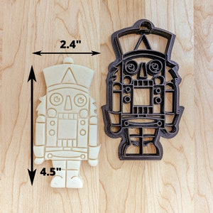 May include: A white cookie cutter in the shape of a nutcracker, measuring 4.5 inches tall and 2.4 inches wide. The cookie cutter is next to a black metal nutcracker cookie cutter of the same design.