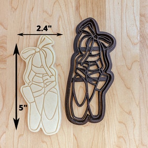 May include: A pair of ballet shoe cookie cutters, one is black and the other is white. The black cutter is 2.4 inches wide and 5 inches tall.