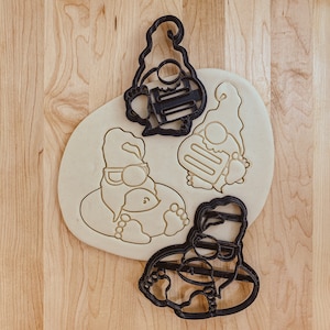 May include: Three black cookie cutters shaped like gnomes. The gnomes are wearing hats and have beards. The cookie cutters are on a wooden surface.