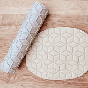 May include: A light gray rolling pin and a piece of dough, both with a repeating geometric cube pattern. The rolling pin is cylindrical and lies next to the oval-shaped dough, which has the same embossed design. The items are on a wooden surface.