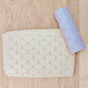 May include: A light blue rolling pin with an anchor pattern next to a piece of dough imprinted with the same anchor design. The dough is beige and sits on a light wood surface. The rolling pin is cylindrical.