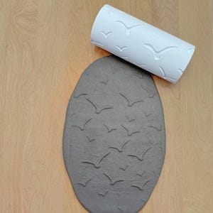 May include: A white rolling pin with a pattern of birds imprinted on a grey clay surface.