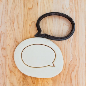 May include: A white cookie cutter in the shape of a speech bubble, with a black plastic speech bubble cookie cutter on top of it.
