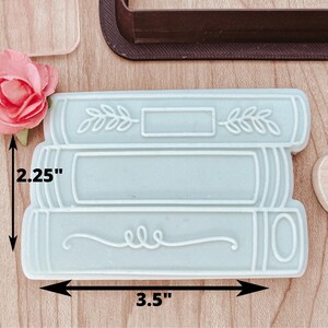 May include: A light blue silicone cookie cutter shaped like a stack of three books. The cutter measures 3.5 inches wide and 2.25 inches tall.
