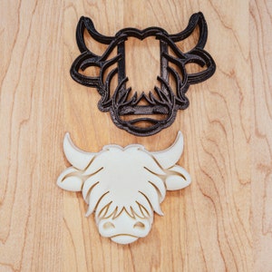 May include: A black and a white Highland cow cookie cutter set. The black cutter is the outline, and the white one is the shape. The cutters are on a light wood grain surface.