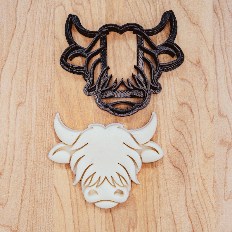 May include: A black and a white Highland cow cookie cutter set. The black cutter is the outline, and the white one is the shape. The cutters are on a light wood grain surface.