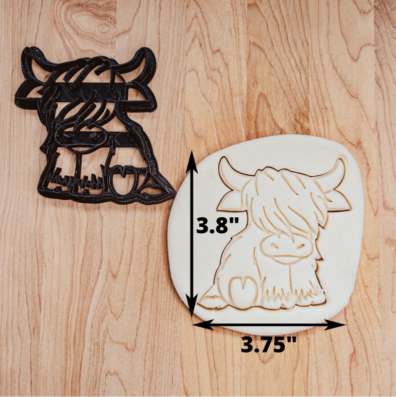 A black, plastic cookie cutter of a cartoon highland cow. The imprint made by the cutter are shown in dough beside the cutter. The cow is sitting with it's tail and one back foot visible. The fur on top of it's head covers both eyes.