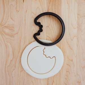 May include: A black plastic cookie cutter shaped like a crescent moon with a bite taken out of it, on top of a white fondant cookie dough.