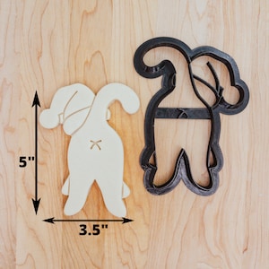 May include: A cookie cutter and a cut-out biscuit in the shape of a gnome's backside. The cookie cutter is black, and the biscuit is beige. The gnome's backside has a Santa hat and a stitched design. The biscuit is 12.7 cm tall and 8.9 cm wide.