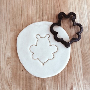 May include: A white cookie dough with a bee-shaped cookie cutter pressed into it. The cookie cutter is a black plastic bee shape.