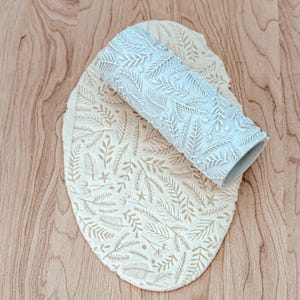 May include: A white rolling pin with a leaf and star pattern is rolling over a piece of dough with the same pattern. The dough is on a wooden surface.