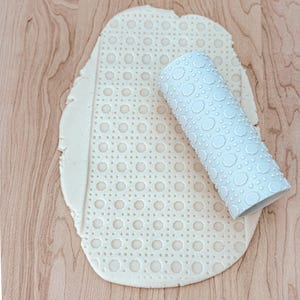 May include: A white rolling pin with a geometric pattern is used to create a patterned design on a sheet of dough.