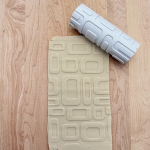 May include: A white rolling pin with a geometric pattern is rolling over a beige sheet of dough with the same pattern embossed into it.