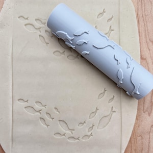 May include: A gray rolling pin with a fish design is used to create an imprint of fish on a sheet of dough.
