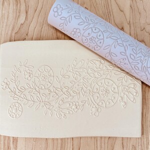 May include: A light pink rolling pin with a floral design lies on a rectangular piece of clay with a matching embossed floral pattern. The rolling pin is cylindrical, and the clay is off-white.