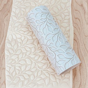 May include: A white rolling pin with a leaf pattern design. The rolling pin is used to create an embossed leaf pattern on dough.