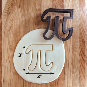 Peut inclure: Un emporte-pièce en plastique noir en forme de symbole mathématique pi, de 7,6 cm de diamètre. L'emporte-pièce est présenté sur une surface en bois avec une pâte blanche.