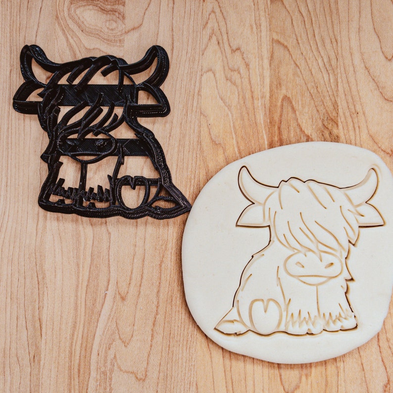 Puede incluir: Un cortador de galletas de pl&aacute;stico negro en forma de vaca de las tierras altas, con una impresi&oacute;n de masa de galletas blanca del mismo dise&ntilde;o.