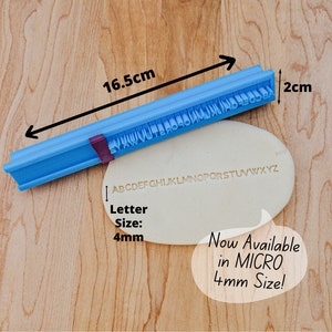 May include: A blue plastic alphabet stamp with a purple handle. The stamp is 16.5 cm long and 2 cm wide. The letters are 4 mm tall. The stamp is shown on a white surface with the alphabet printed in the surface.