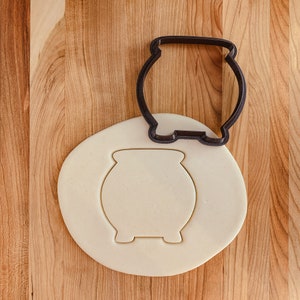May include: A black plastic cookie cutter in the shape of a cauldron on a white surface. The cookie cutter is on top of a round piece of white cookie dough.