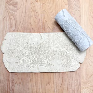 May include: A light gray clay roller and a rectangular piece of clay with a maple leaf pattern. The roller is cylindrical and features a detailed leaf design. The clay piece shows an embossed leaf pattern, suggesting a craft or art project.