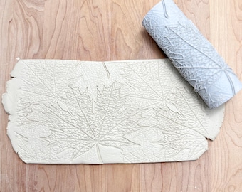 May include: A light gray clay roller and a rectangular piece of clay with a maple leaf pattern. The roller is cylindrical and features a detailed leaf design. The clay piece shows an embossed leaf pattern, suggesting a craft or art project.