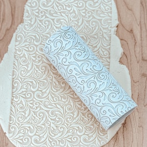 Peut inclure: Rouleau à pâtisserie blanc avec un motif floral décoratif, roulant sur une feuille de pâte blanche avec le même motif.