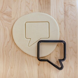 May include: A close-up of a cookie cutter in the shape of a speech bubble. The cookie cutter is made of metal and has a black finish. The cookie cutter is sitting on a wooden surface.