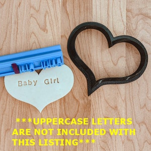 May include: A blue and white plastic cookie cutter in the shape of a heart. The cookie cutter is on a wooden surface. A white heart-shaped cookie is next to the cutter. The cookie has the words "Baby Girl" stamped on it. The text "***UPPERCASE LETTERS ARE NOT INCLUDED WITH THIS LISTING***" is written in yellow on the wooden surface.