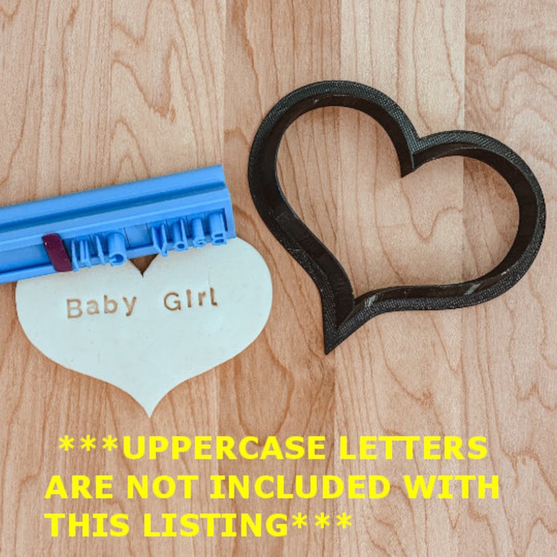 May include: A blue and white plastic cookie cutter in the shape of a heart. The cookie cutter is on a wooden surface. A white heart-shaped cookie is next to the cutter. The cookie has the words "Baby Girl" stamped on it. The text "***UPPERCASE LETTERS ARE NOT INCLUDED WITH THIS LISTING***" is written in yellow on the wooden surface.