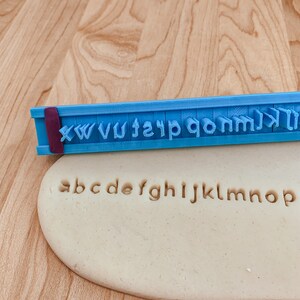 May include: A blue plastic alphabet stamp with the letters X, W, V, U, T, S, R, Q, P, O, N, M, L, K, J, I, H, G, F, E, D, C, B, A. The stamp is used to imprint the letters into a beige play dough.