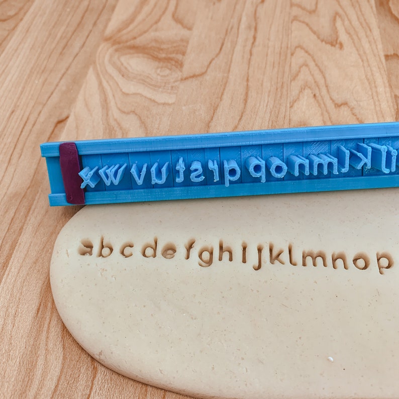 May include: A blue plastic alphabet stamp with the letters X, W, V, U, T, S, R, Q, P, O, N, M, L, K, J, I, H, G, F, E, D, C, B, A. The stamp is used to imprint the letters into a beige play dough.