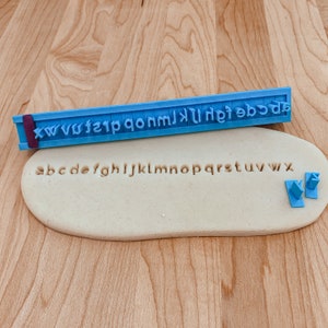 May include: A blue plastic alphabet stamp with the letters xwvwfzip qonm jklhfe dcba. The stamp is used to imprint letters into a beige fondant surface. The letters abcdefghijklmnopqrstuvwxyz are imprinted into the fondant.