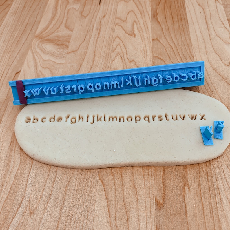 May include: A blue plastic alphabet stamp with the letters xwvwfzip qonm jklhfe dcba. The stamp is used to imprint letters into a beige fondant surface. The letters abcdefghijklmnopqrstuvwxyz are imprinted into the fondant.