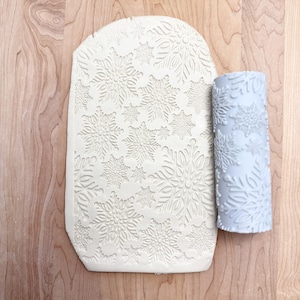 May include: A light grey rolling pin and a rectangular piece of clay, both embossed with a snowflake pattern. The clay is pressed with the snowflake design. The rolling pin is cylindrical and has the same snowflake design.