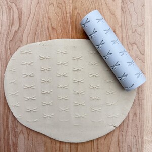 May include: A grey rolling pin with a dragonfly pattern is used to create an embossed design on a piece of beige clay.