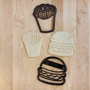 May include: Four cookie cutters, two black and two white, shaped like a hamburger, french fries, and a cup of fries. The cookie cutters are on a light brown wooden surface.