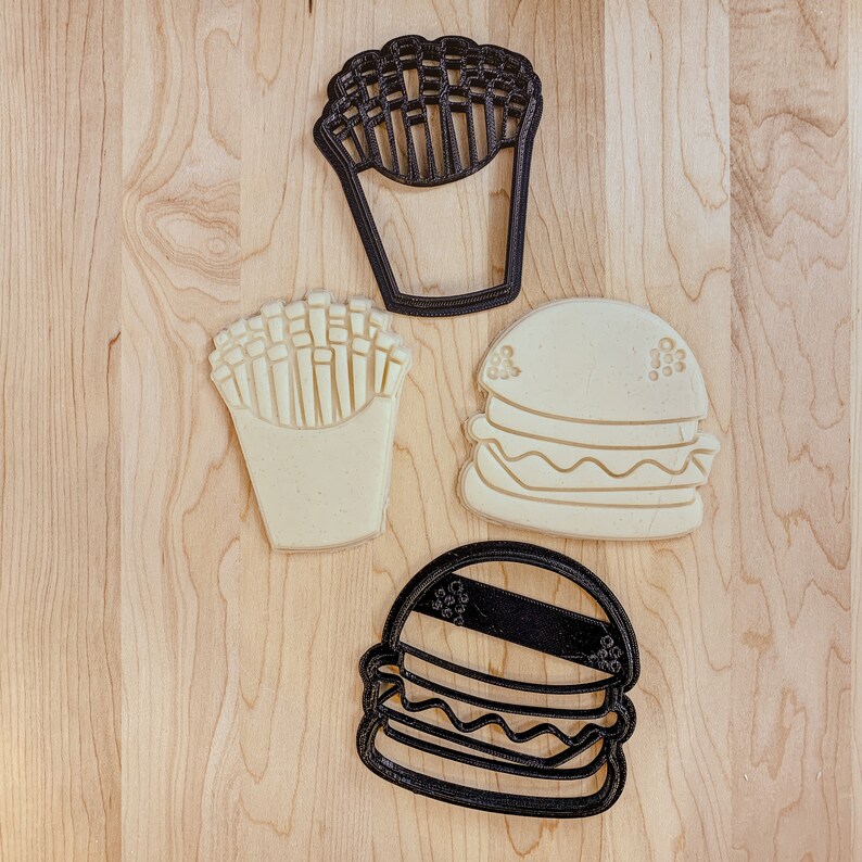 May include: Four cookie cutters, two black and two white, shaped like a hamburger, french fries, and a cup of fries. The cookie cutters are on a light brown wooden surface.