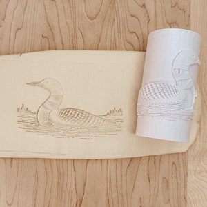 May include: A light-colored, carved stamp and a matching cylindrical roller featuring a detailed illustration of a loon swimming in water. The loon is depicted with intricate feather and water details, creating a textured effect.