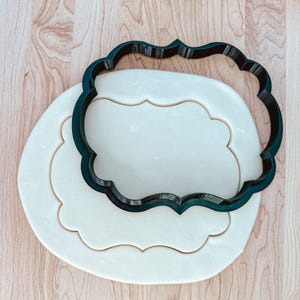 May include: A black plastic cookie cutter in the shape of a decorative frame, placed on top of a white fondant cookie dough.