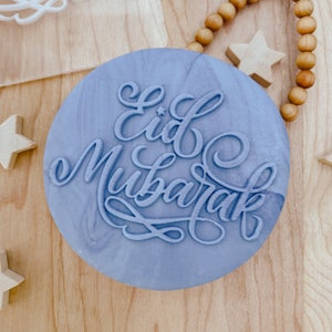 May include: A light blue round stamp with the words "Eid Mubarak" in a decorative script.