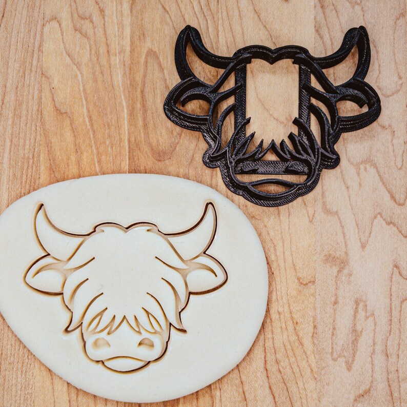 May include: A black plastic Highland cow cookie cutter next to a cookie with the cow's face imprinted. The cookie cutter is in the shape of the cow's head with horns and shaggy fur. The cookie is round and has the cow's face pressed into it.