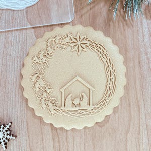 May include: A beige cookie cutter with a detailed design of a Christmas wreath with holly leaves, a star, and a nativity scene. The cookie cutter is on a wooden surface.
