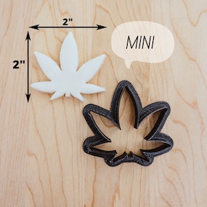 May include: A white plastic 2-inch cannabis leaf cookie cutter and a black 2-inch cannabis leaf cookie cutter on a wooden surface. The text "MINI" is written in a speech bubble.