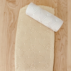 May include: A white, patterned rolling pin rests on a piece of clay with a matching circular design. The clay is light brown, and the rolling pin has a repeating swirl pattern. The background is a light wood surface.